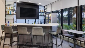Un área de bar moderna con un televisor grande, taburetes y estanterías llenas de botellas y copas.