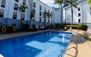 Una piscina con borde de baldosas, rodeada de palmeras y un edificio con varias ventanas.
