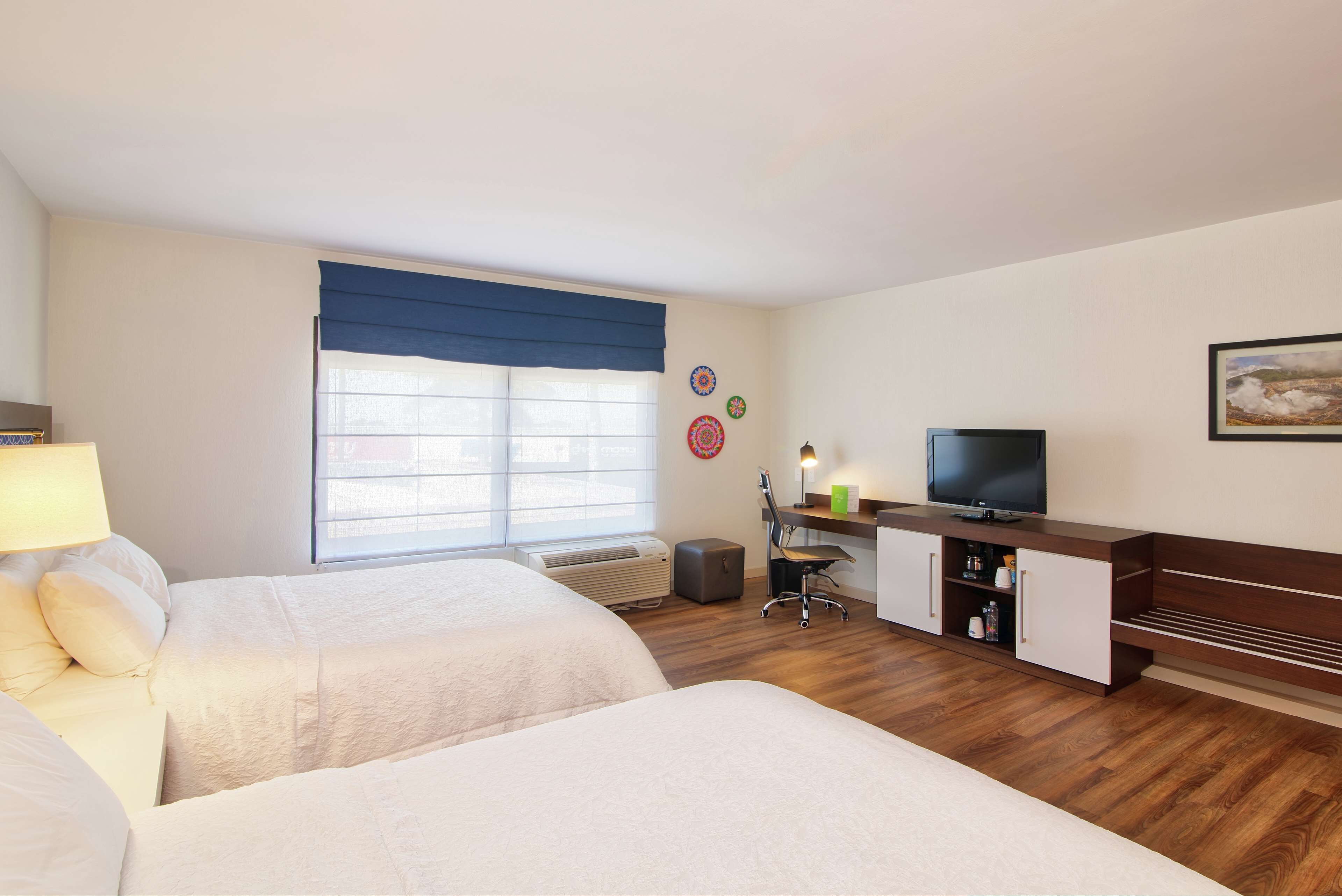 A hotel room with two beds, a desk with a chair, a television, and a window with a blue curtain.