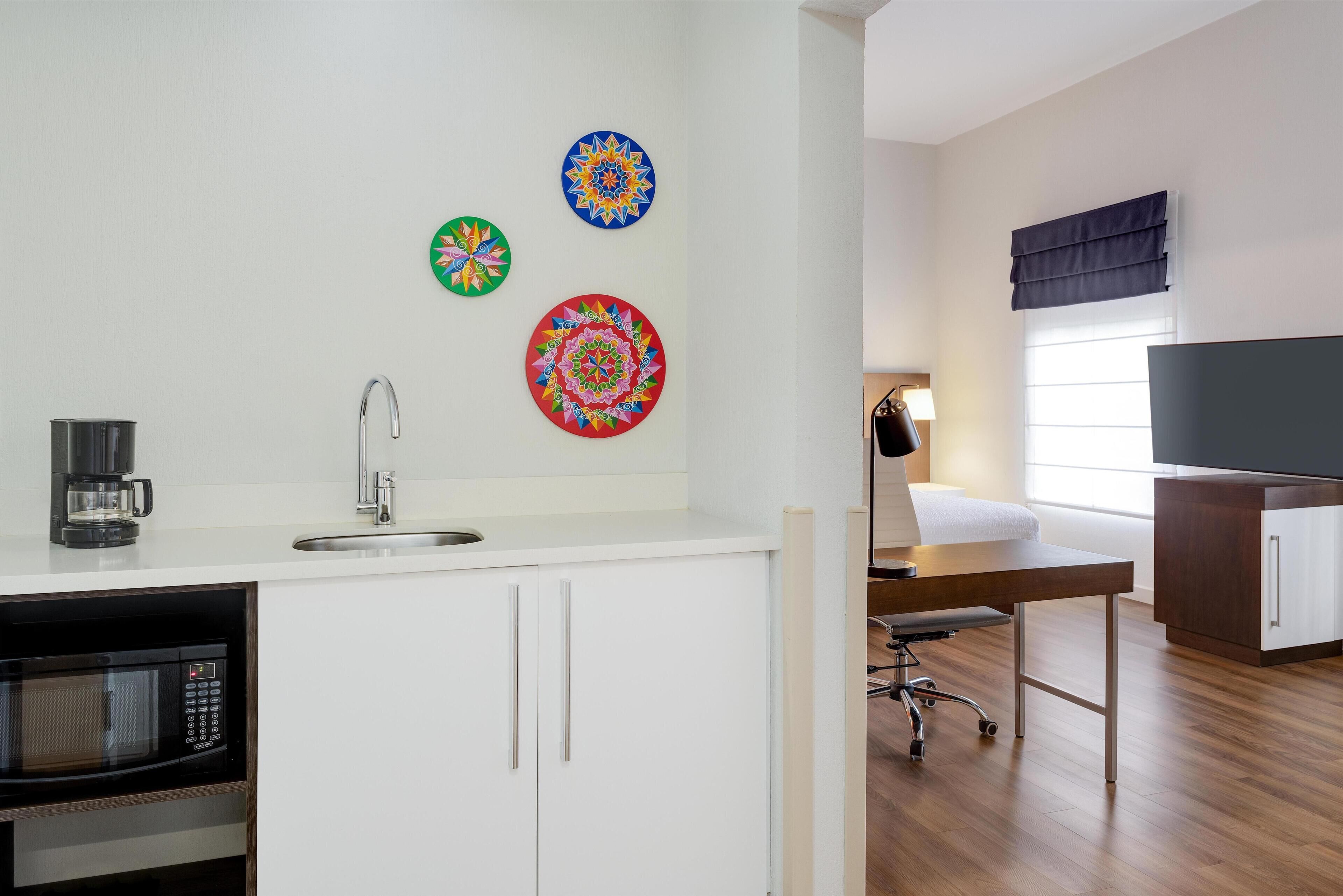 A modern kitchen with a microwave, sink, and coffee maker. There are colorful wall decorations and a TV in the adjacent room.