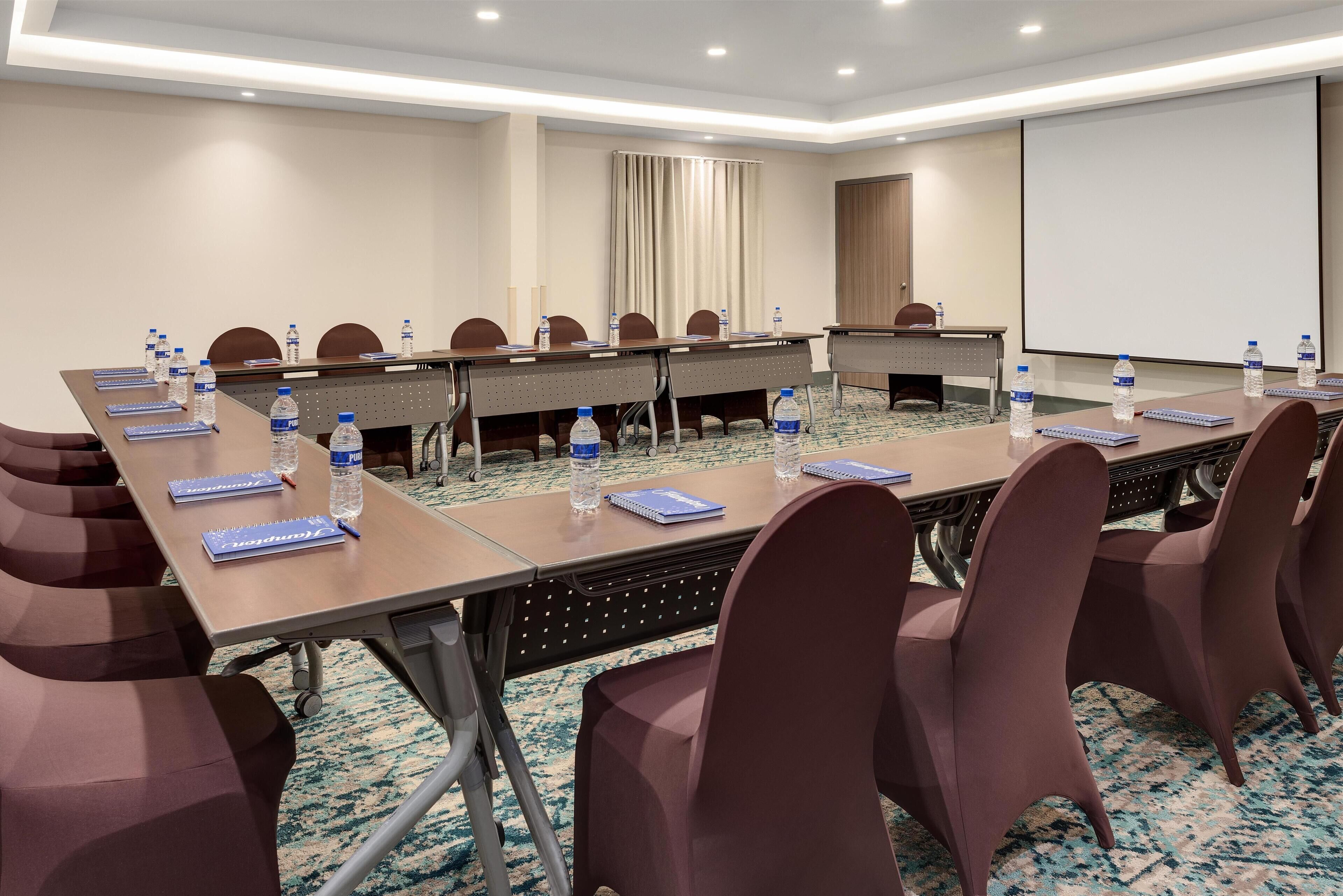 A conference room with a long table, chairs, water bottles, notebooks, and a projector screen.