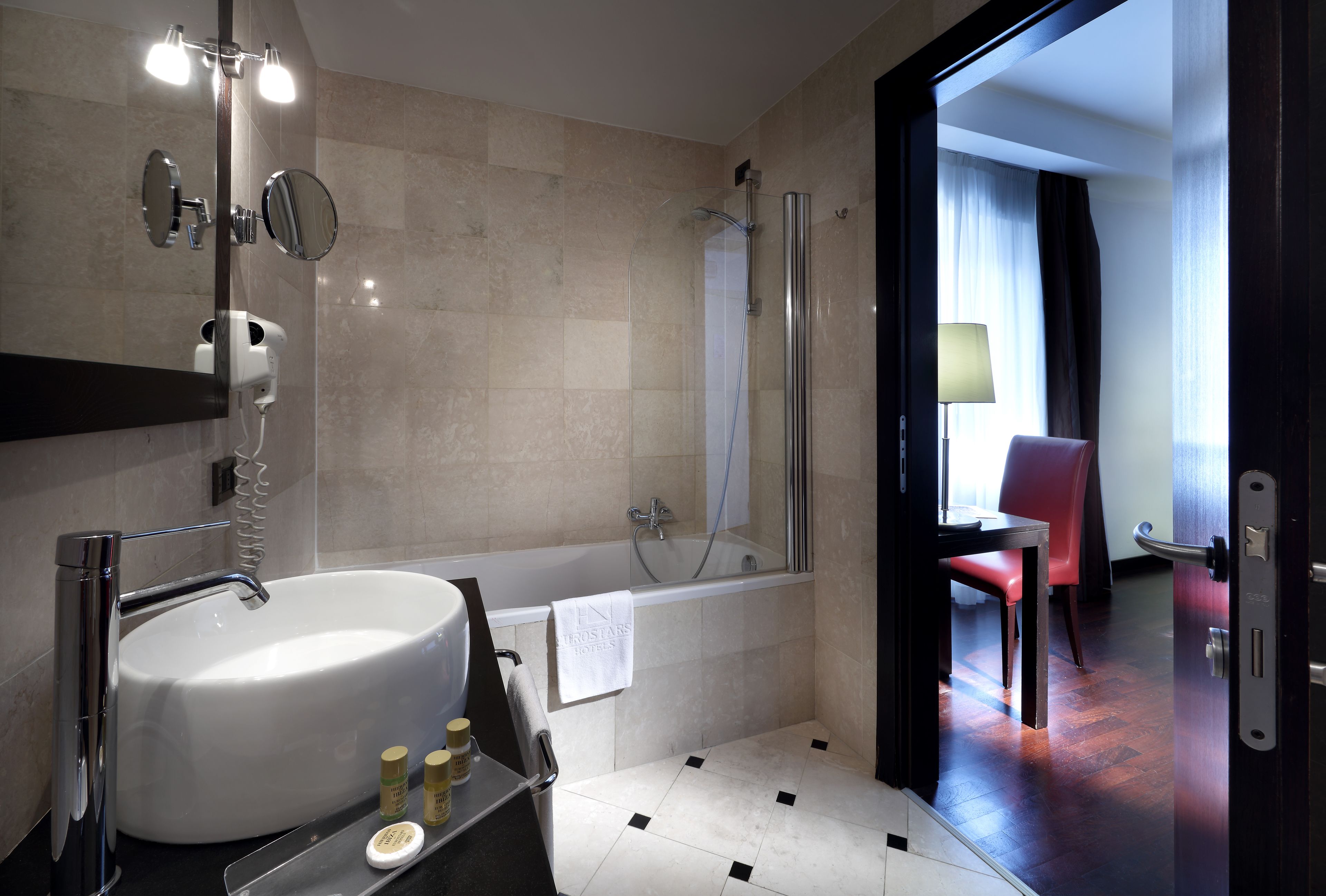 A modern bathroom with a large white sink, a glass-enclosed shower, and a mirror with lights. A towel hangs on the wall, and a red chair is visible in the adjacent room.