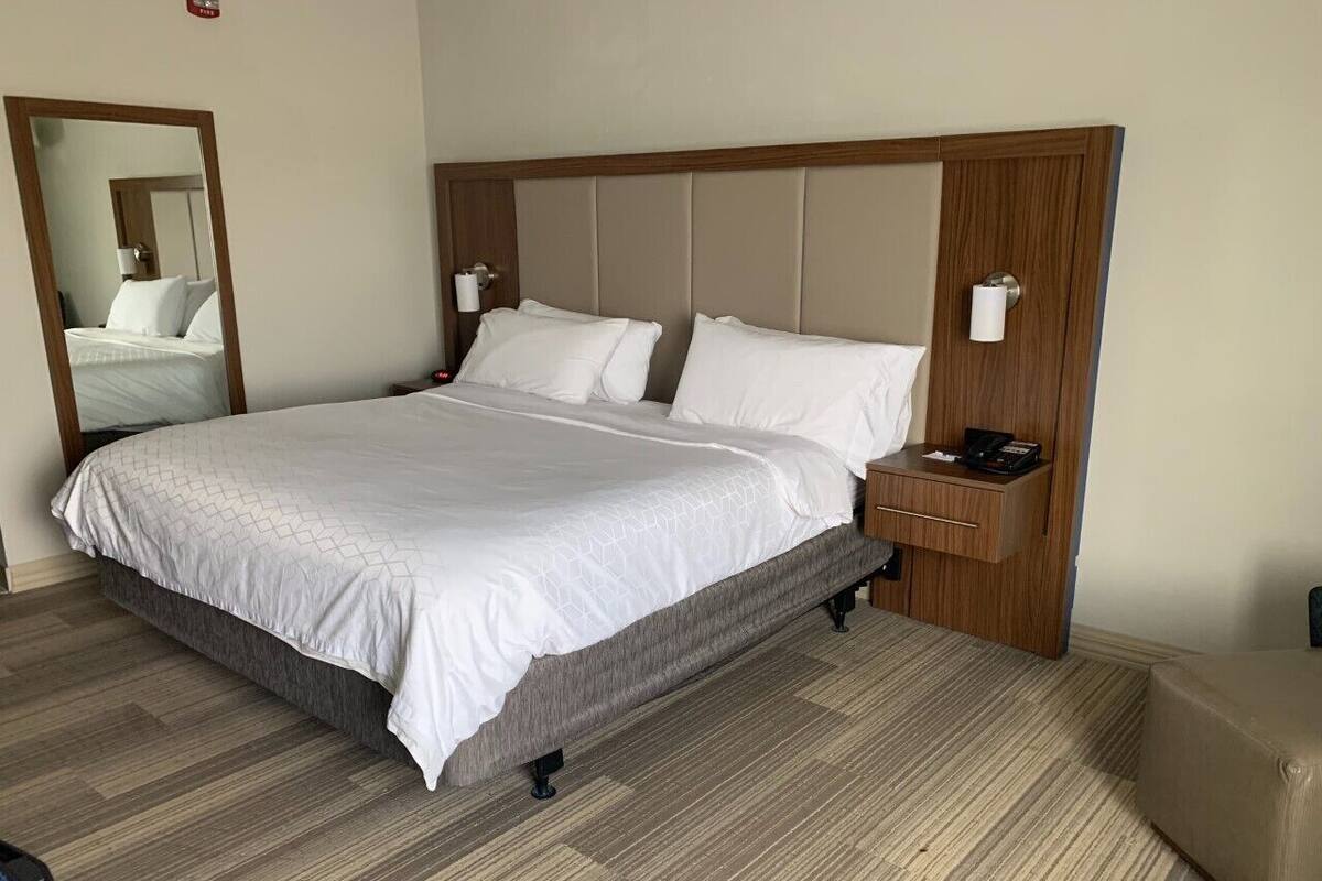 Una cama con cuidada presentación, cabecera de madera, mesita de noche con lámpara y un espejo grande en la pared.