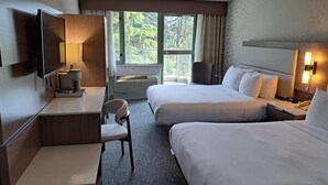 Habitación de hotel con dos camas, un escritorio con lámpara, una silla, un televisor y una ventana con vistas a zonas verdes.