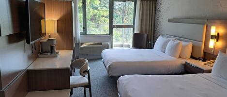 Camera d'albergo con due letti, una scrivania con una lampada, una sedia, una TV e una finestra con vista sul verde.