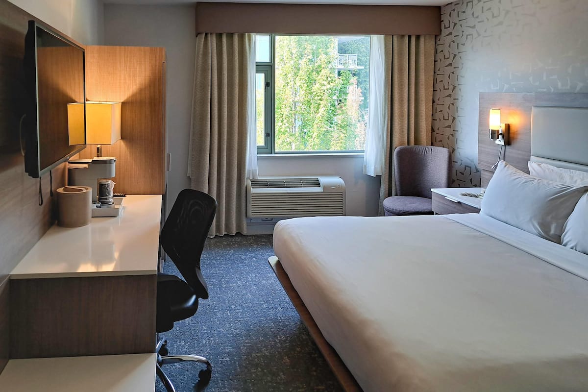 Une chambre d’hôtel comprenant un lit, un bureau avec une lampe, une chaise, une fenêtre avec des rideaux, un climatiseur et un luminaire fixé au mur.