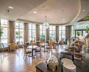 Un vaste espace dédié à la restauration, avec des tables et des chaises rondes, de grandes fenêtres et un buffet proposant une variété de plats.