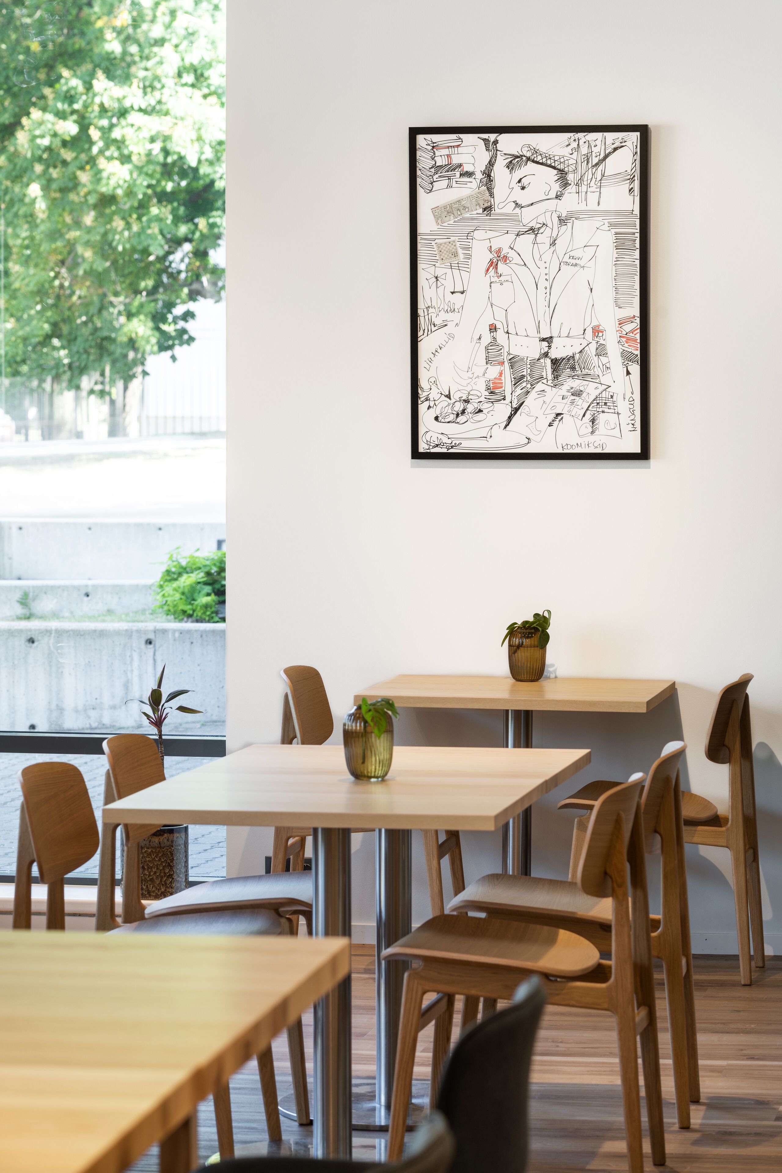 Un espace repas avec des tables et des chaises en bois, une illustration en noir et blanc encadrée au mur et une plante en pot posée sur l’une des tables.
