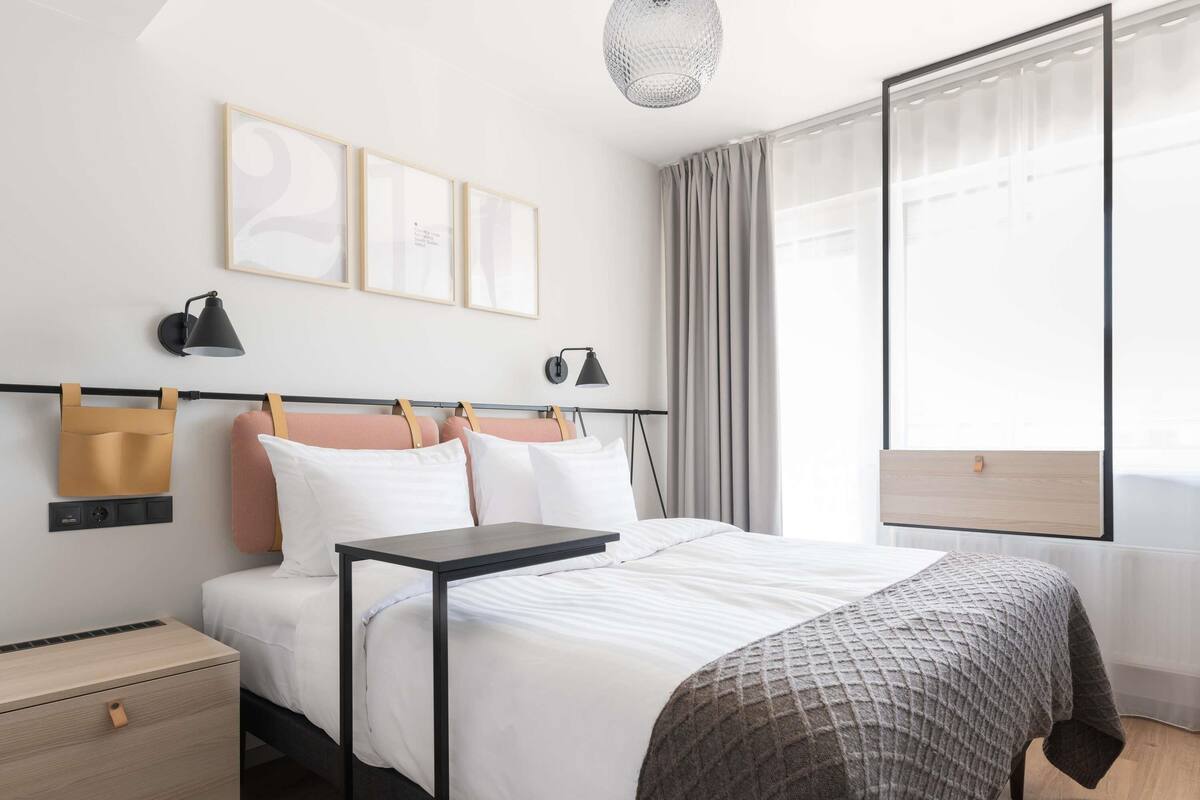 A modern hotel room with a bed, bedside tables, a desk, and a large window with curtains.