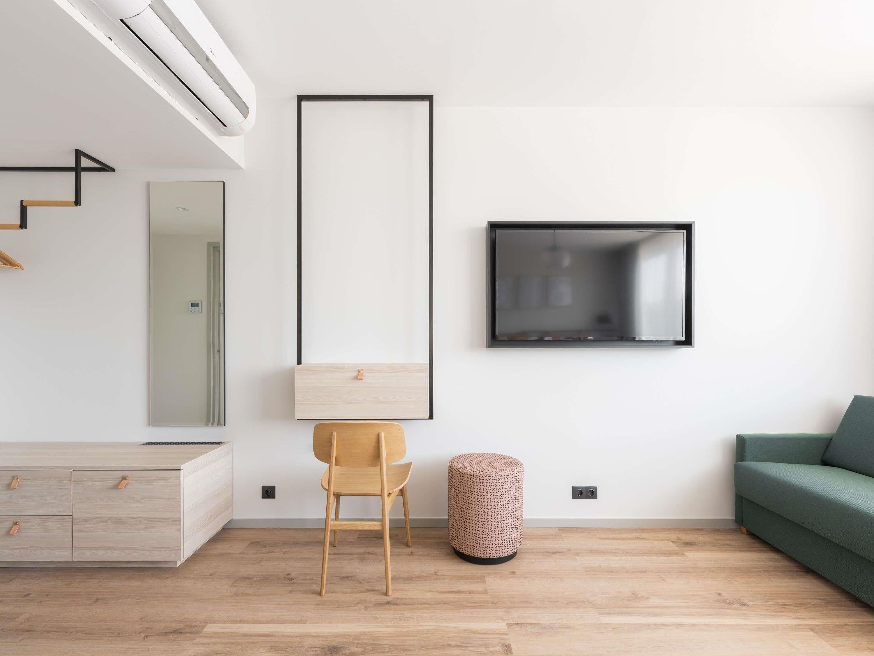 Ein modernes Wohnzimmer mit Holzboden, einem grünen Sofa, einem Holzstuhl, einem kleinen runden Hocker, einem an der Wand befestigten Fernseher und einem minimalistischen Design.