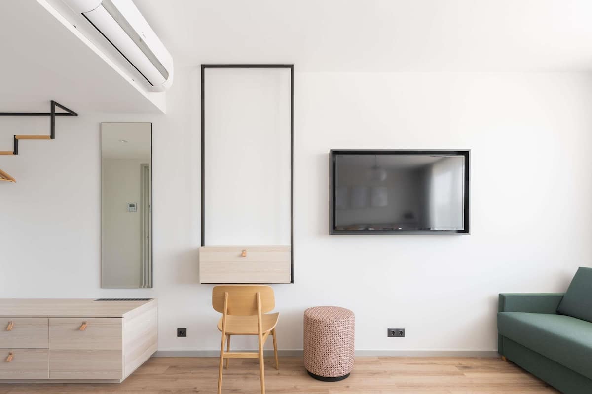 Ein modernes Wohnzimmer mit Holzboden, einem grĂŒnen Sofa, einem Holzstuhl, einem kleinen runden Hocker, einem an der Wand befestigten Fernseher und einem minimalistischen Design.