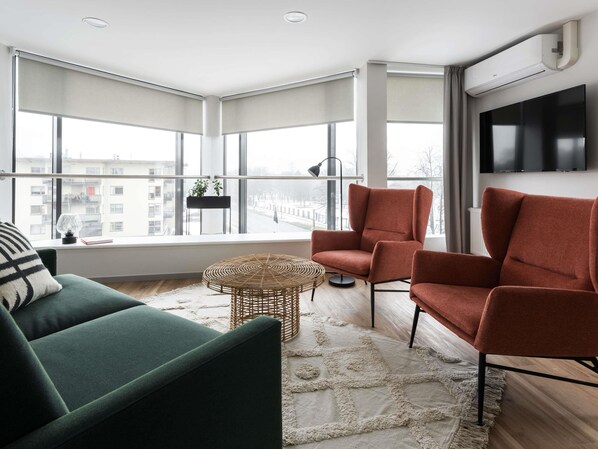 A modern living room with a green sofa, two red armchairs, a round coffee table, and a large window with a view of buildings.