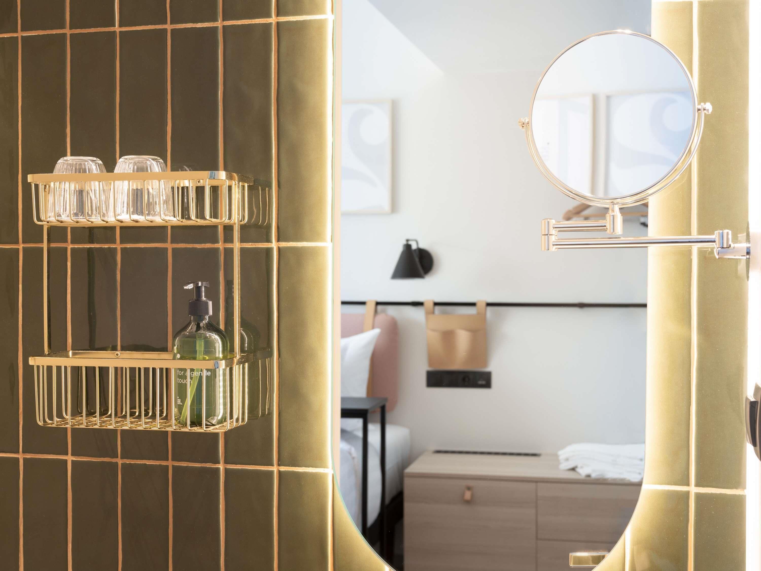 Habitación de hotel con cama, mesita de noche, lámpara, espejo y estante con artículos de aseo.