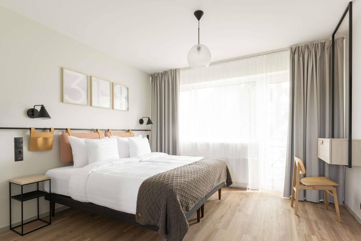 Une chambre d’hôtel moderne, dotée d’un grand lit, de tables de chevet, d’un bureau, d’une chaise et d’une grande fenêtre avec des rideaux.