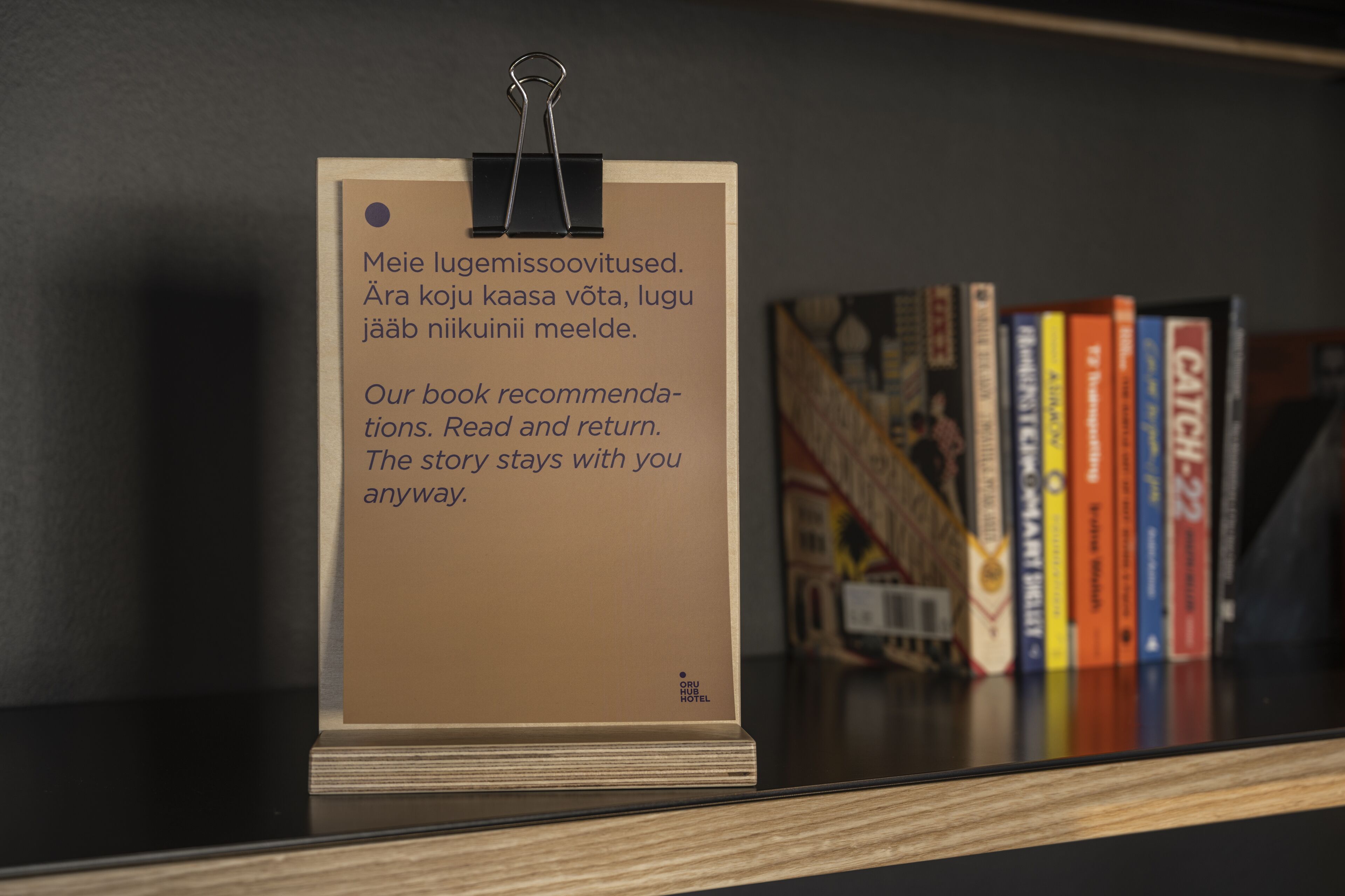 Ein Buchtipp-Schild mit Holzfuß und Metallclip, platziert auf einem Regal mit einer Reihe Bücher im Hintergrund.