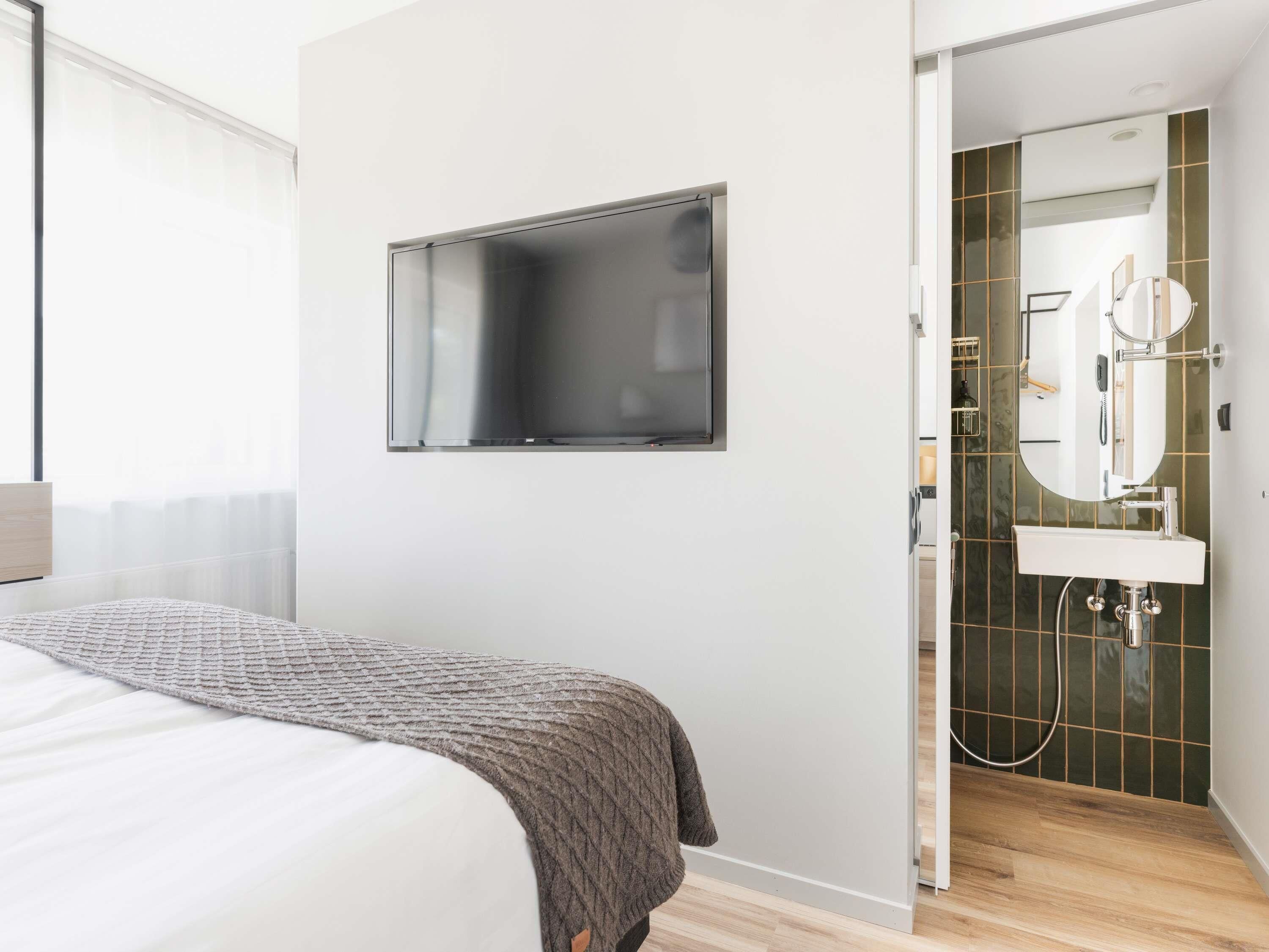Ein modernes Schlafzimmer mit Bett, einem an der Wand befestigten Fernseher und einem Badezimmer mit Waschbecken und Spiegel.