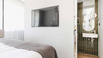 Ein modernes Schlafzimmer mit Bett, einem an der Wand befestigten Fernseher und einem Badezimmer mit Waschbecken und Spiegel.