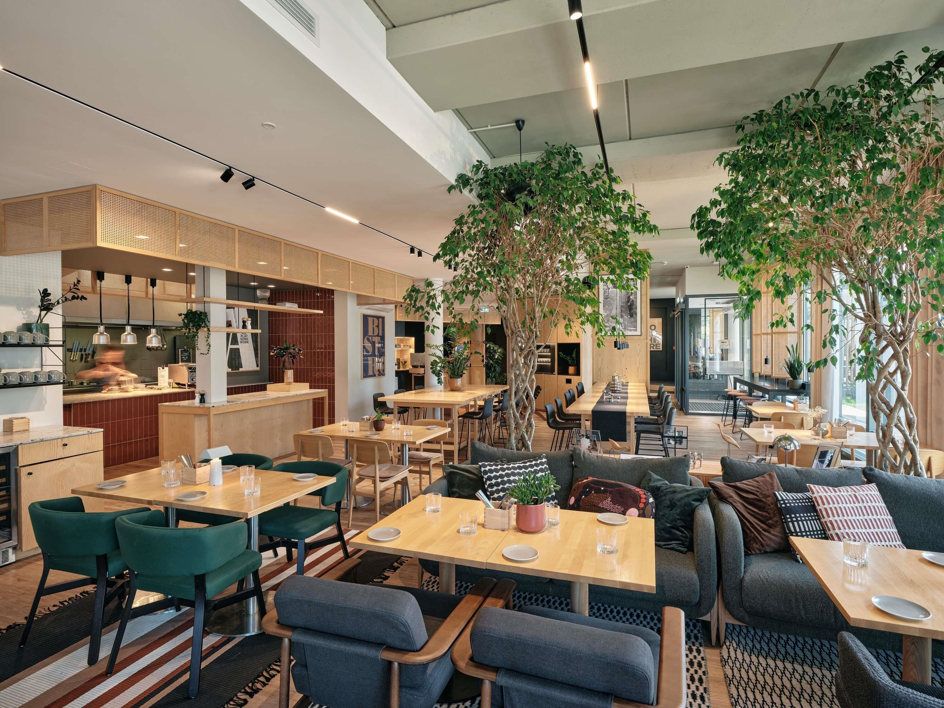 A modern restaurant interior with wooden tables, green chairs, and large potted plants.