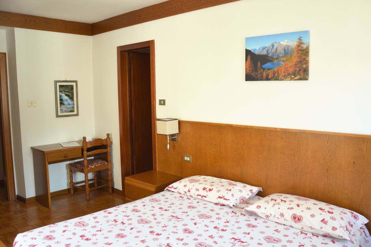 A bedroom with a wooden headboard, a bed with floral bedding, a desk with a chair, a framed picture on the wall, and a painting of a mountain landscape.