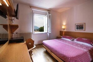 A hotel room with a bed, a desk, a chair, a window with a view, a bedside lamp, and a wall-mounted TV. - Hotel Pinzolo Dolomiti (Pinzolo)