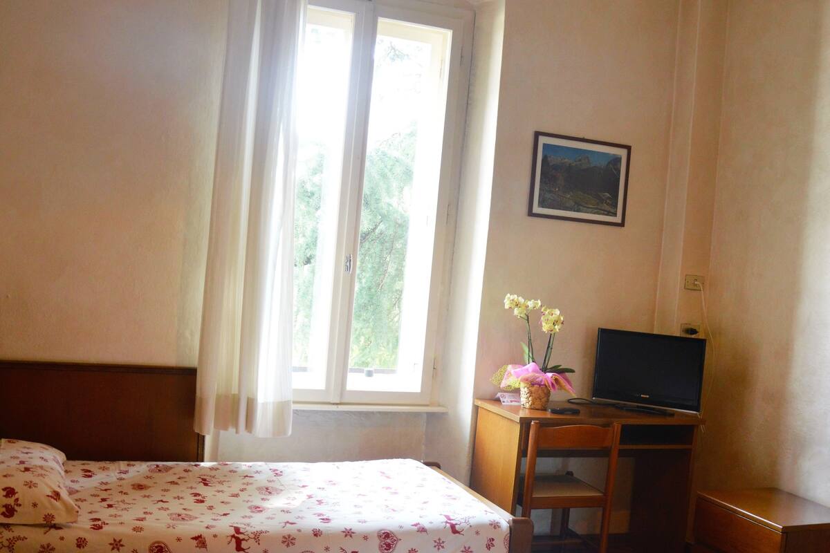 A hotel room with a bed, a desk with a computer, a chair, a lamp, and a window with curtains.