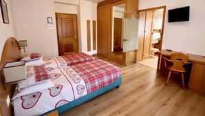 A bedroom with a bed, a desk, and a chair. There is a TV on the wall and a door leading to another room. - Hotel Pinzolo Dolomiti (Pinzolo)