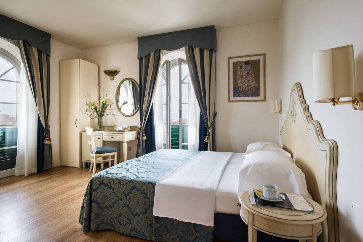 A hotel room with a bed, a desk, a chair, a small table with a cup and a menu, and a window with curtains.