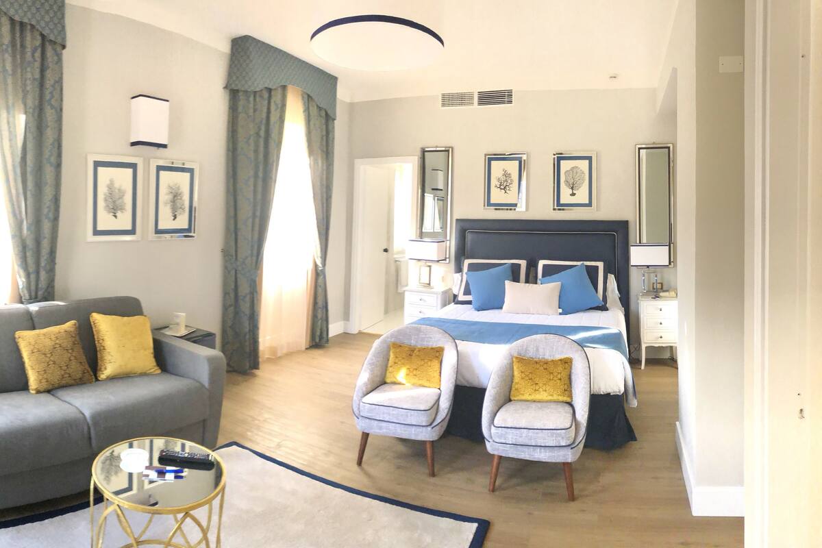A hotel room with a grey sofa, two armchairs, a bed with blue and white bedding, a bedside table, a lamp, and framed pictures on the wall.