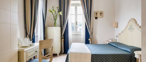 A hotel room with a bed, a desk with a chair, a nightstand, and a window with curtains.