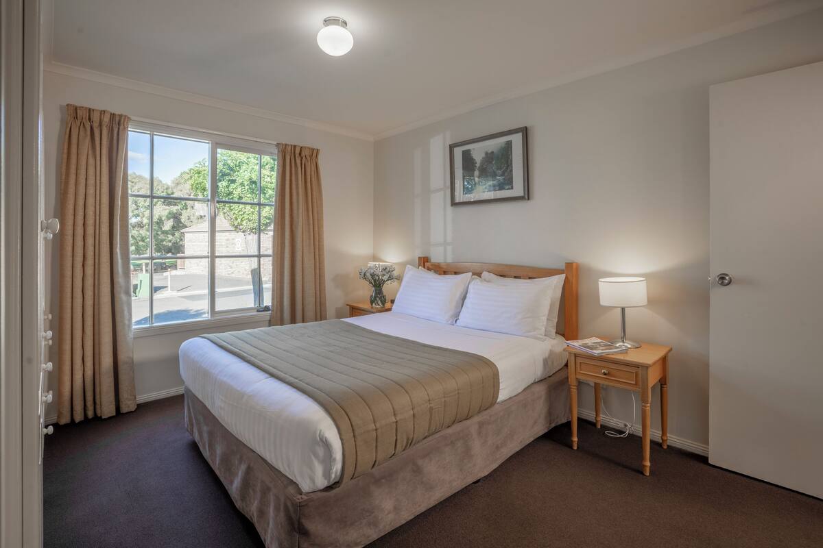Une chambre d’hôtel avec un grand lit, une table de chevet avec une lampe, un tableau encadré au mur et une fenêtre avec des rideaux.