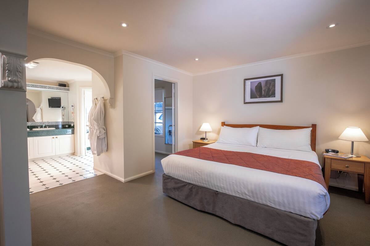 Une chambre d’hôtel avec un grand lit, deux tables de chevet avec des lampes, un tableau encadré au mur et une vue sur la cuisine et la salle de bain, visibles à travers une arche.