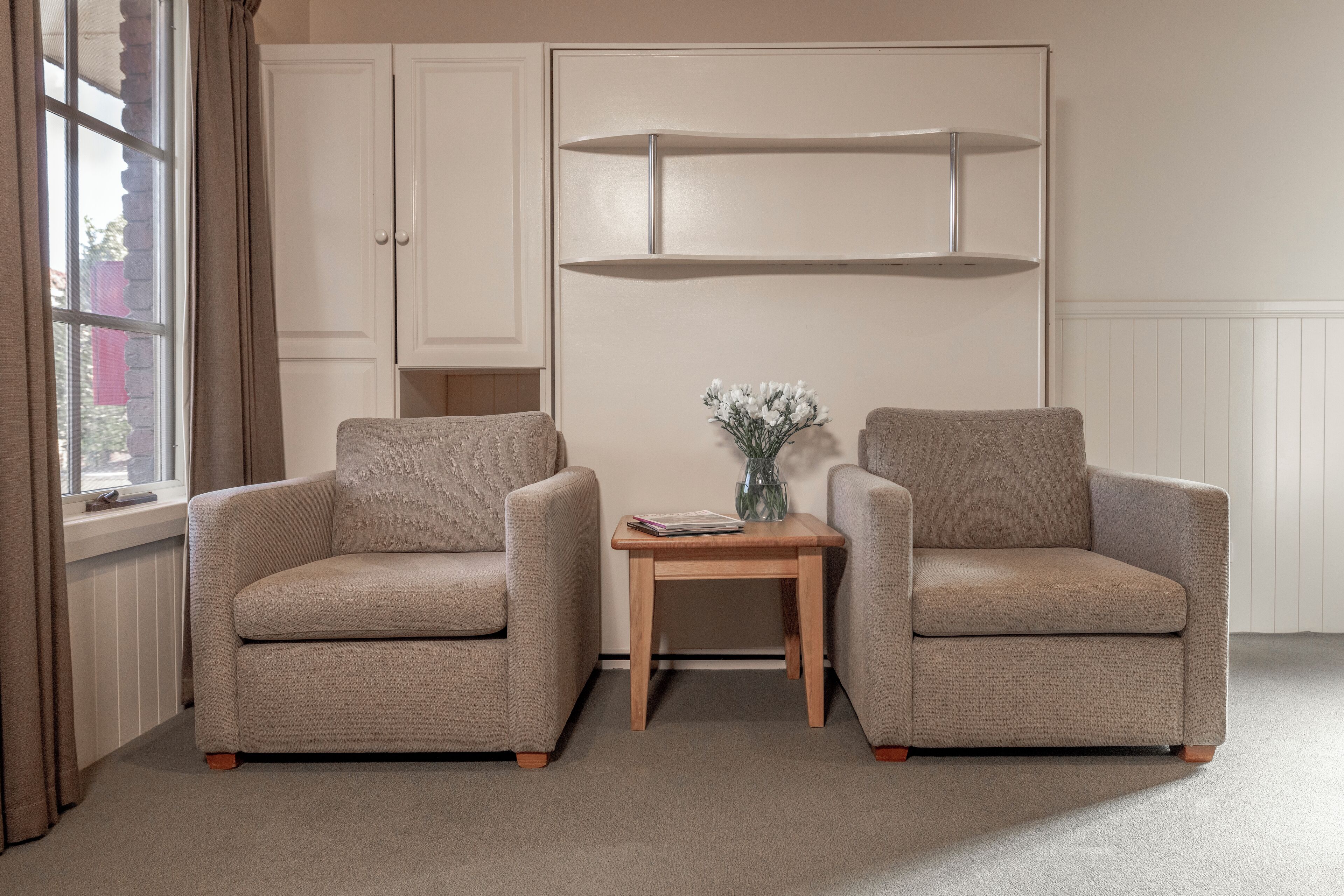 Een kamer met twee beige fauteuils, een houten bijzettafeltje met een vaas en een witte kast.