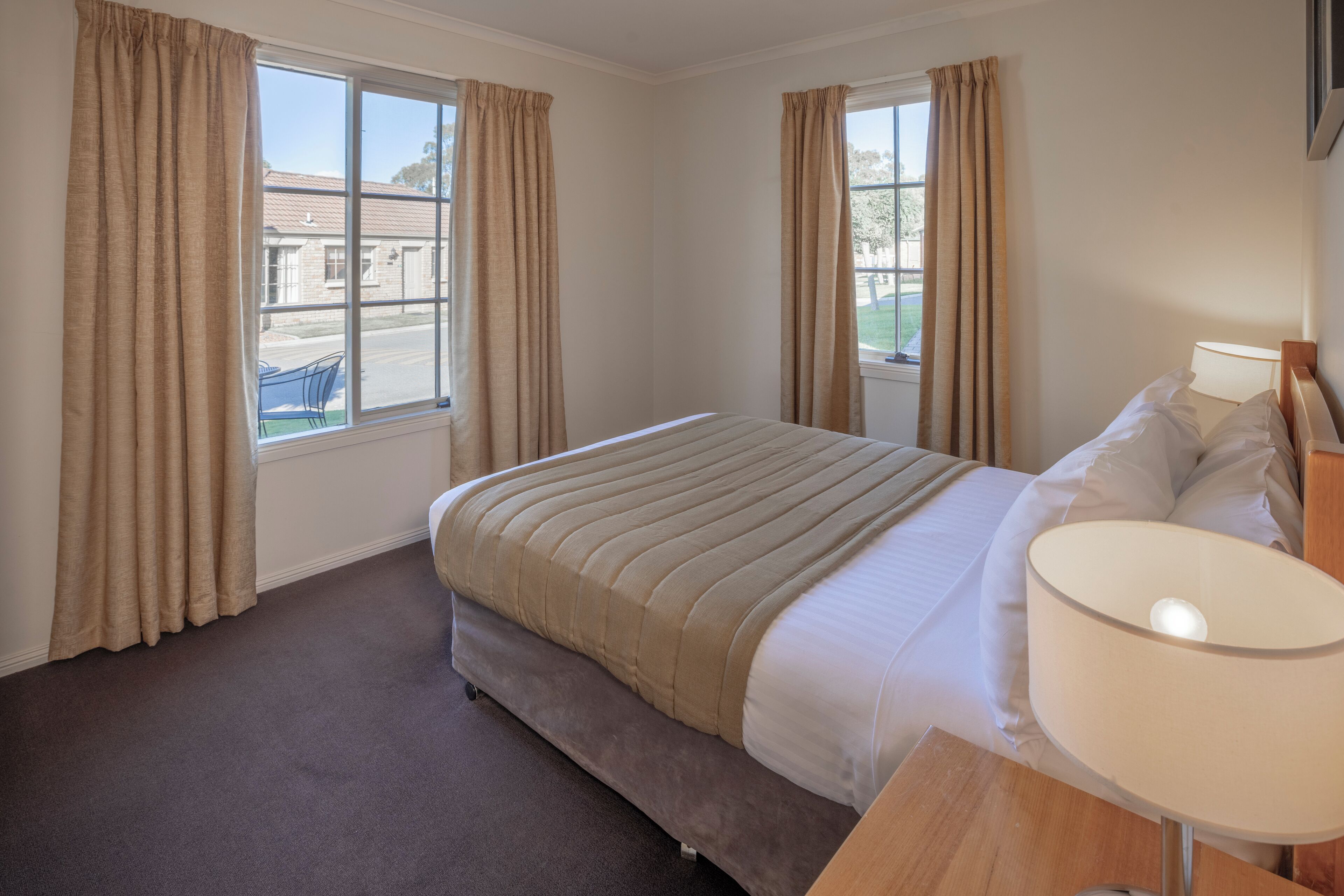 A hotel room with a bed, a bedside table with a lamp, and two windows with curtains.