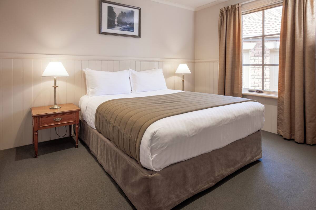 Une chambre d’hôtel bien rangée, avec un grand lit, deux tables de chevet avec des lampes, un tableau encadré au mur et une fenêtre avec des rideaux.