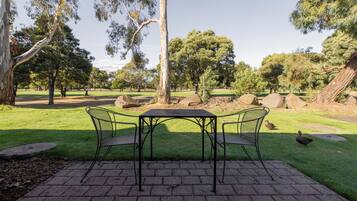 Un patio empedrado con una mesa y sillas de metal, rodeado de césped y árboles.