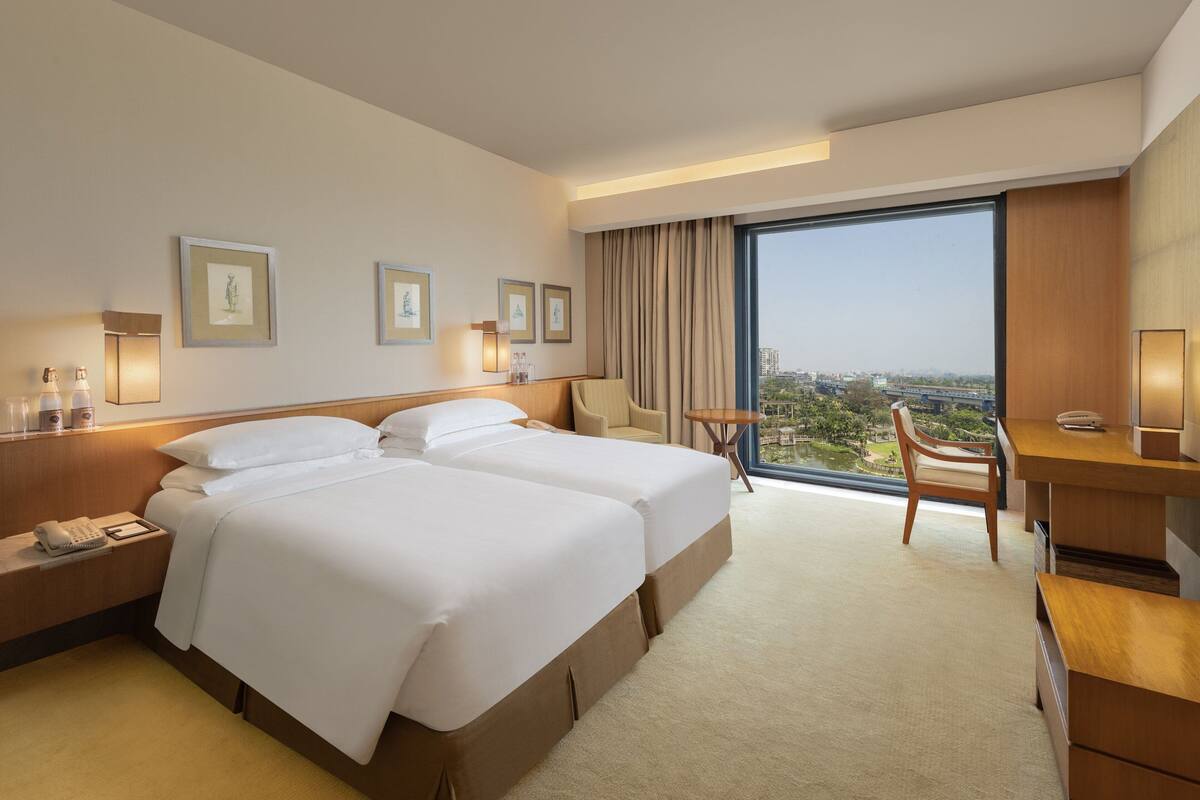 Habitación de hotel con dos camas, un escritorio, una silla y un gran ventanal con vista a la ciudad.