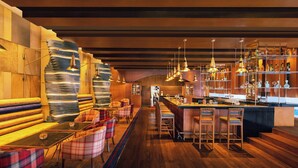 A bar area with wooden flooring, bar stools, and a long counter. There are multiple hanging pendant lights and shelves with bottles.