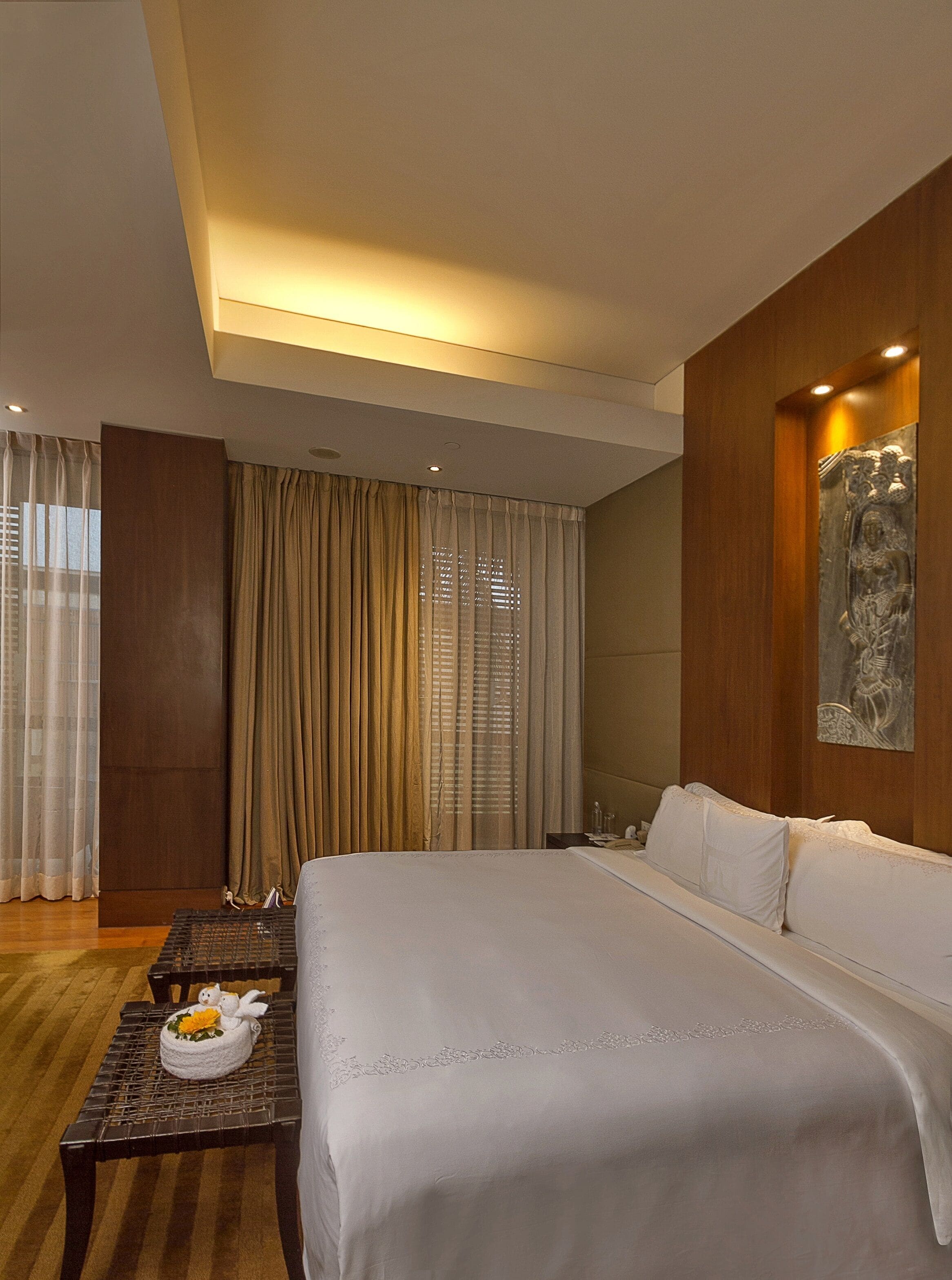 A hotel room with a large bed, a wooden headboard, a small table with a decorative item, and a large window with curtains.