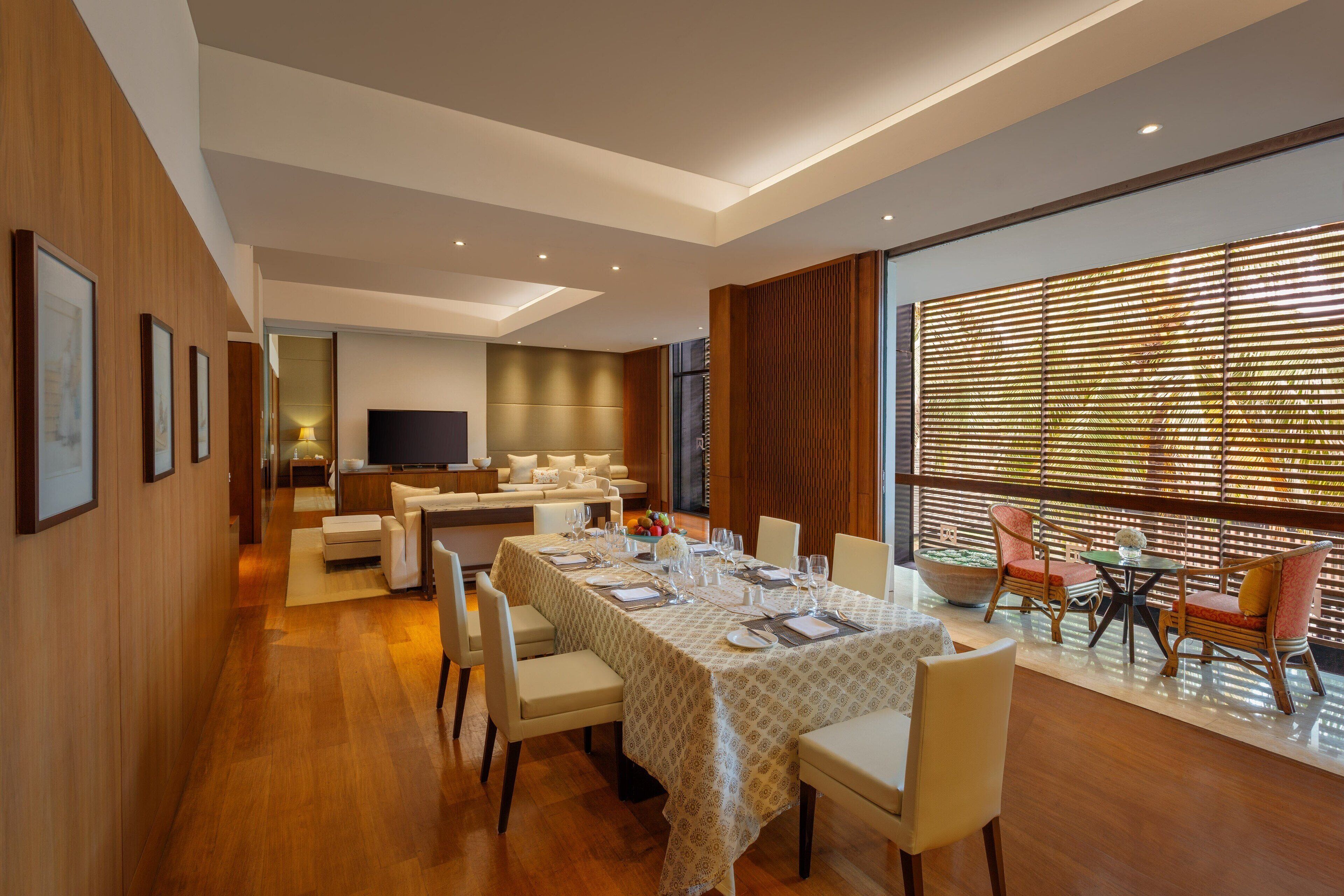 A modern dining area with a long table set for six, surrounded by chairs. A sofa and a television are visible in the background.