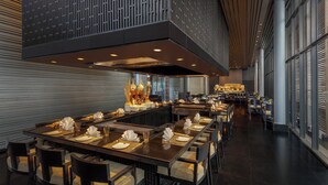 A modern restaurant with a long dining table, multiple chairs, and a high ceiling with exposed beams.