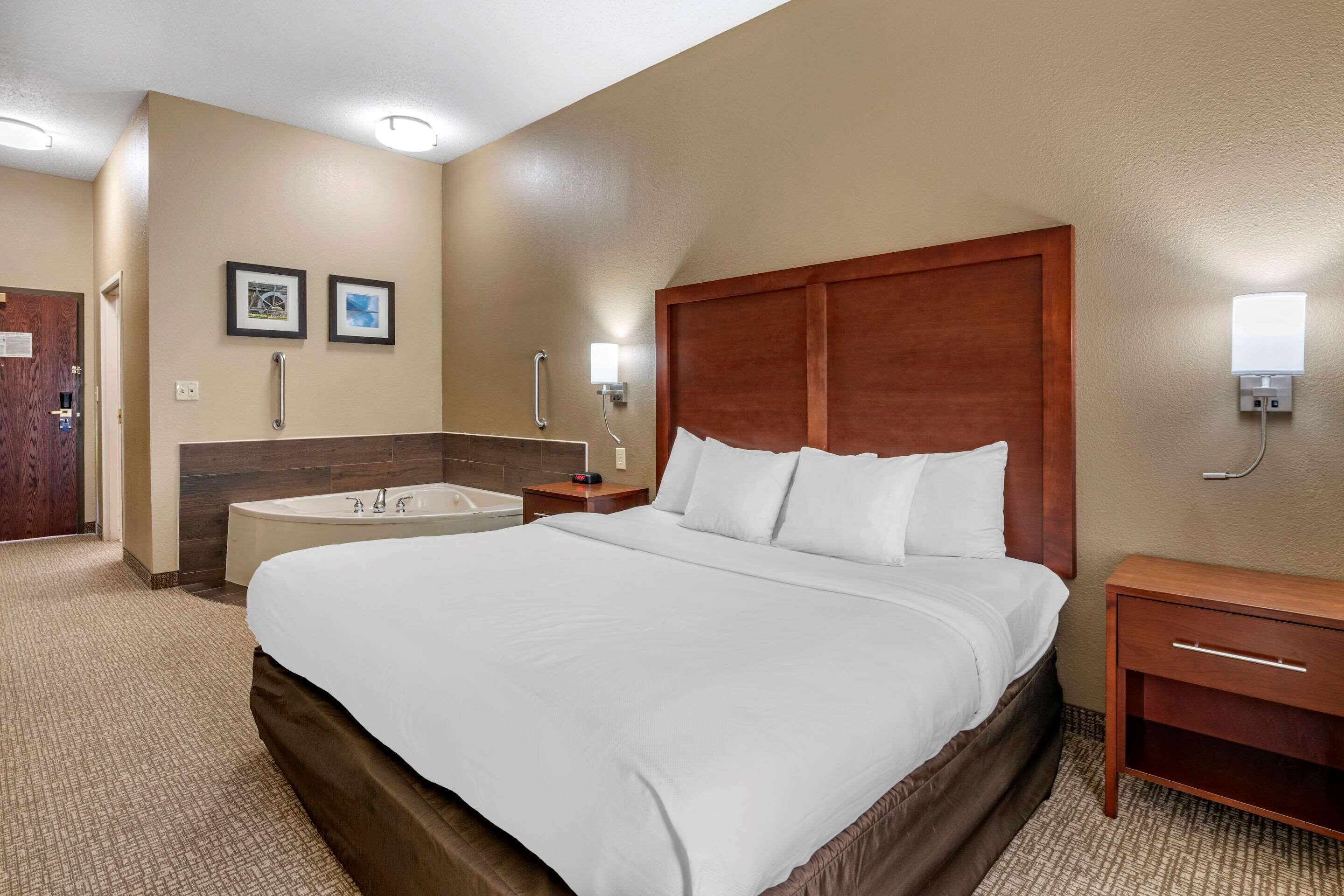A hotel room with a large bed, a wooden headboard, a bedside table with a lamp, a small nightstand, a wall-mounted light, and a bathroom with a bathtub and a towel rack.