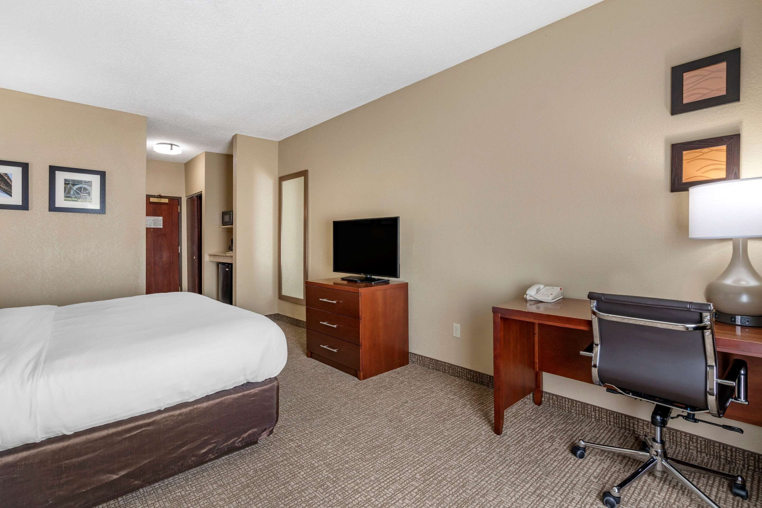 Quarto de hotel com uma cama, uma escrivaninha com cadeira, uma televisão e uma luminária.