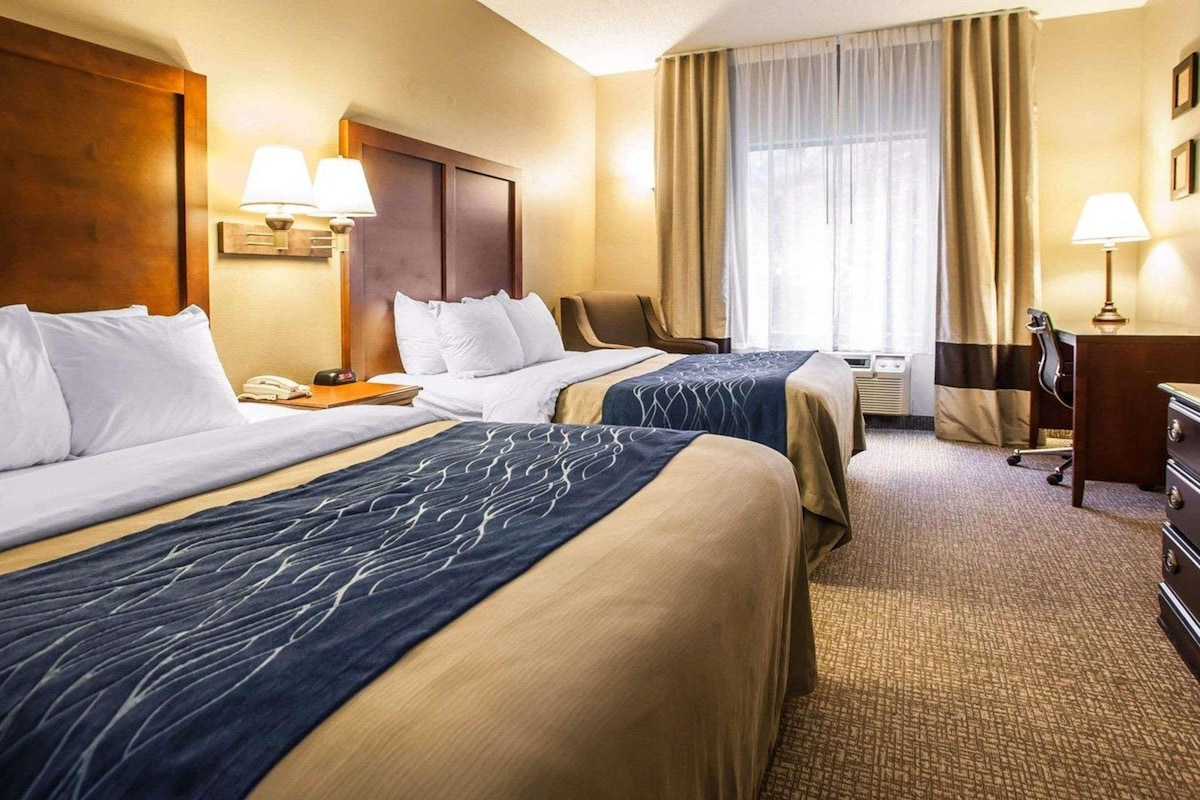 A hotel room with two beds, a desk, a chair, and a dresser. There is a window with curtains and a lamp on the desk.