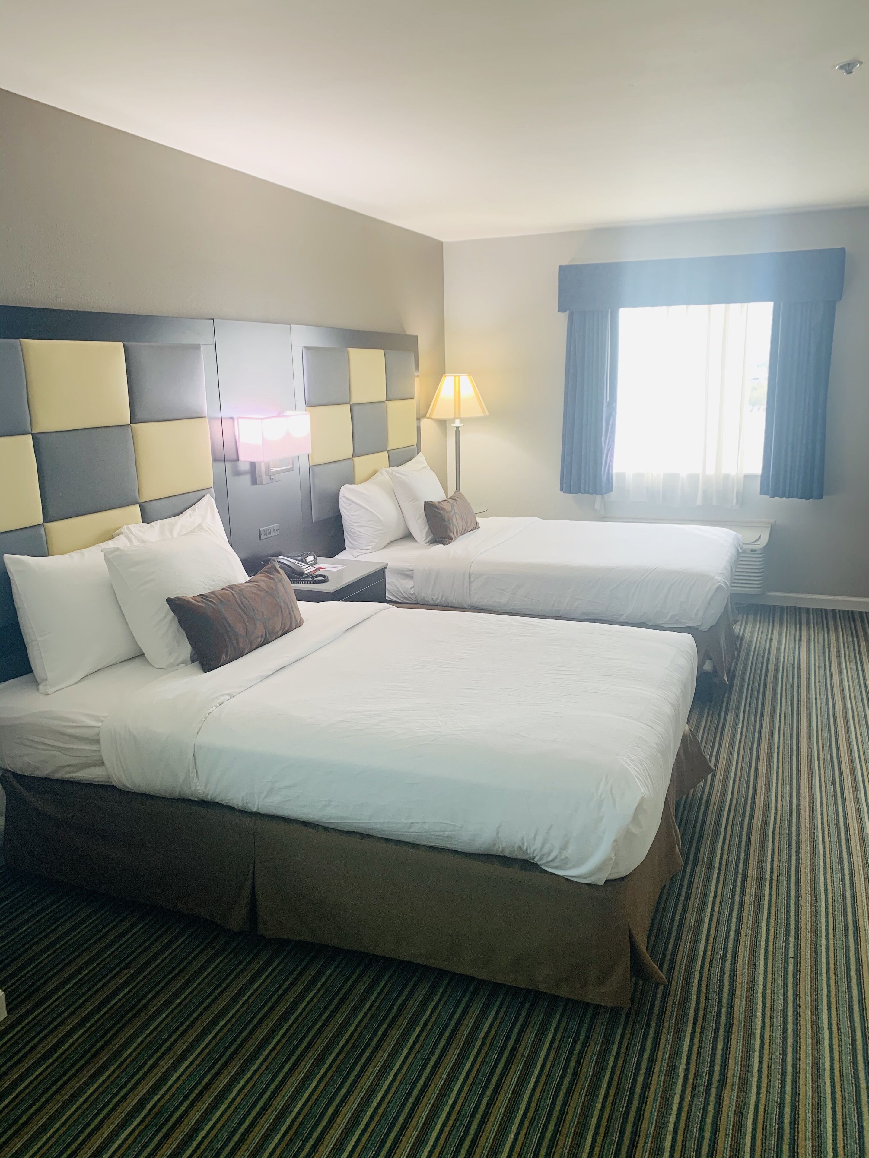 A hotel room with two beds, a headboard with a checkered pattern, a bedside lamp, and a window with curtains.