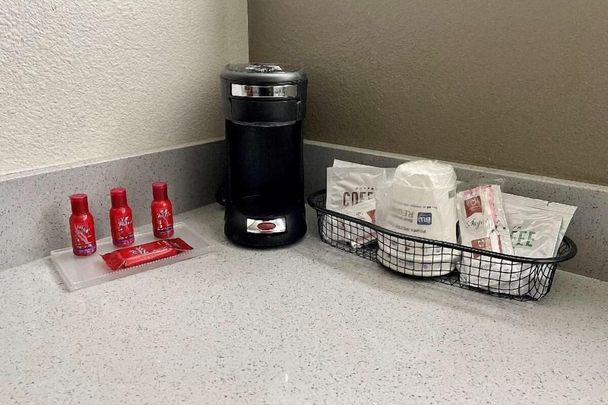 Eine Ecke eines Hotelzimmers mit einem Mülleimer, einem kleinen Tisch mit roten Flaschen und einem Korb mit Taschen und einer Kaffeemaschine.