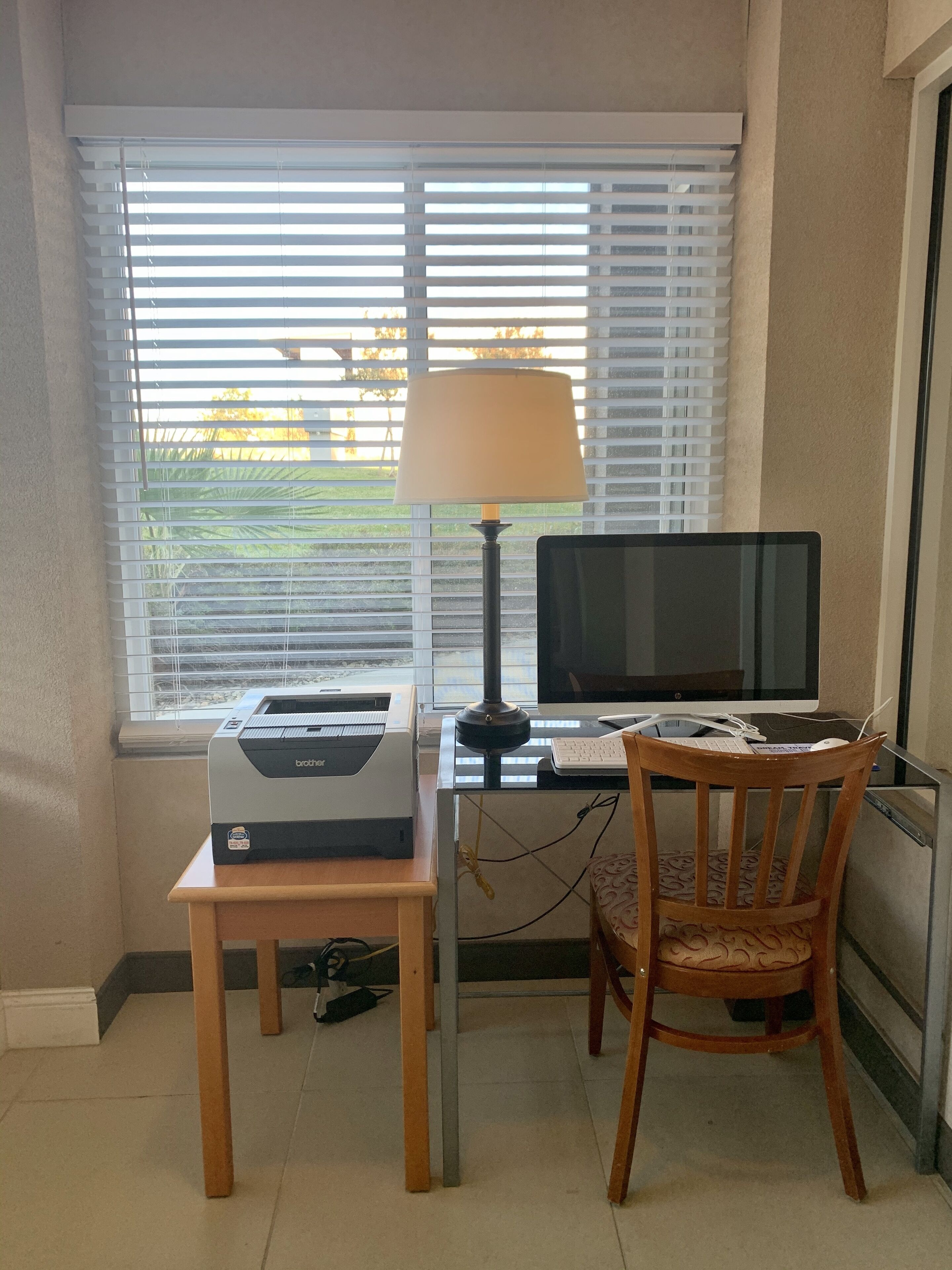 Un espace de travail aménagé à la maison, comprenant une imprimante, un ordinateur et une lampe, près d’une fenêtre munie de stores.