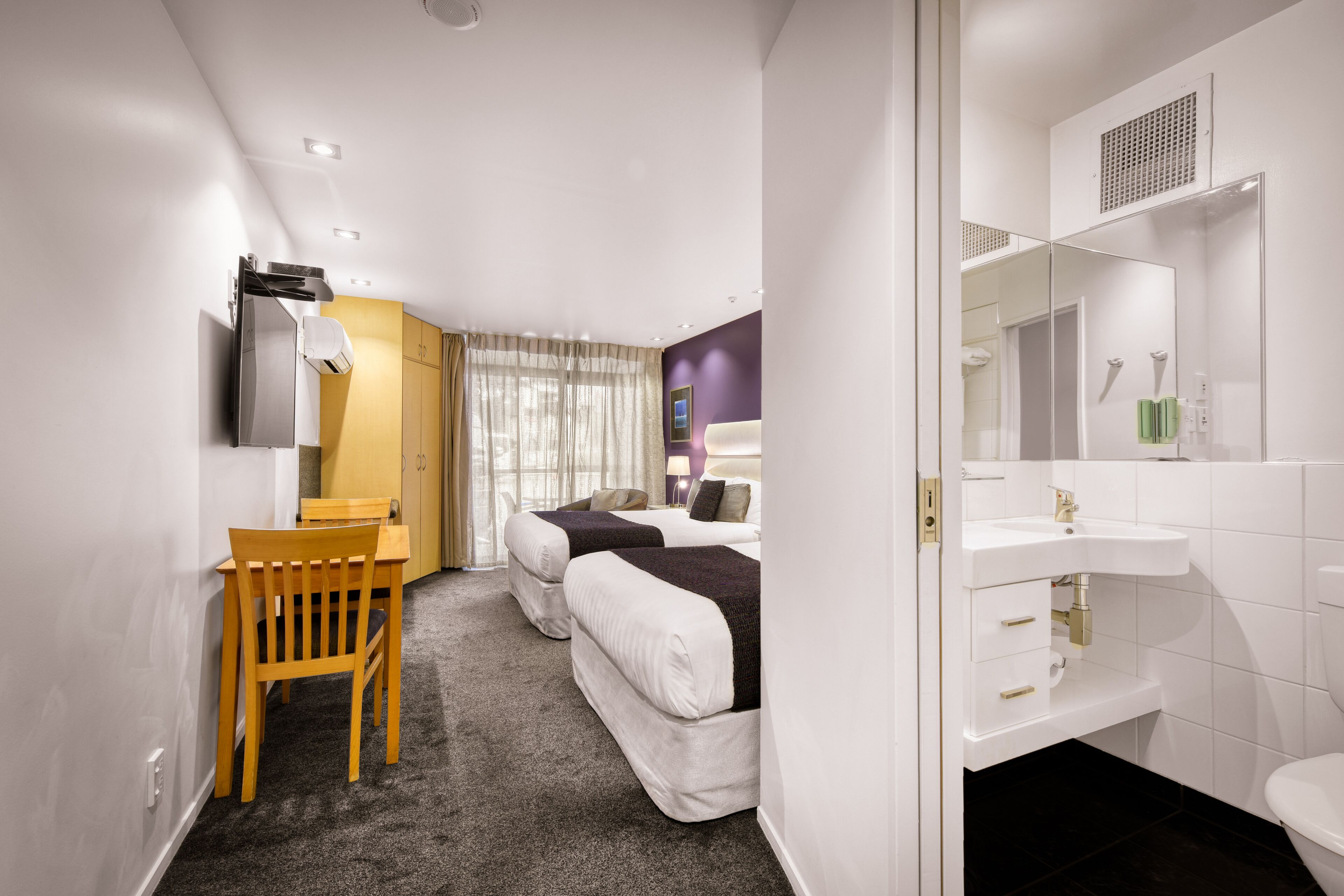 Hotelkamer met twee bedden, een bureau, een stoel, een badkamer met wastafel en spiegel, en een raam met gordijnen.