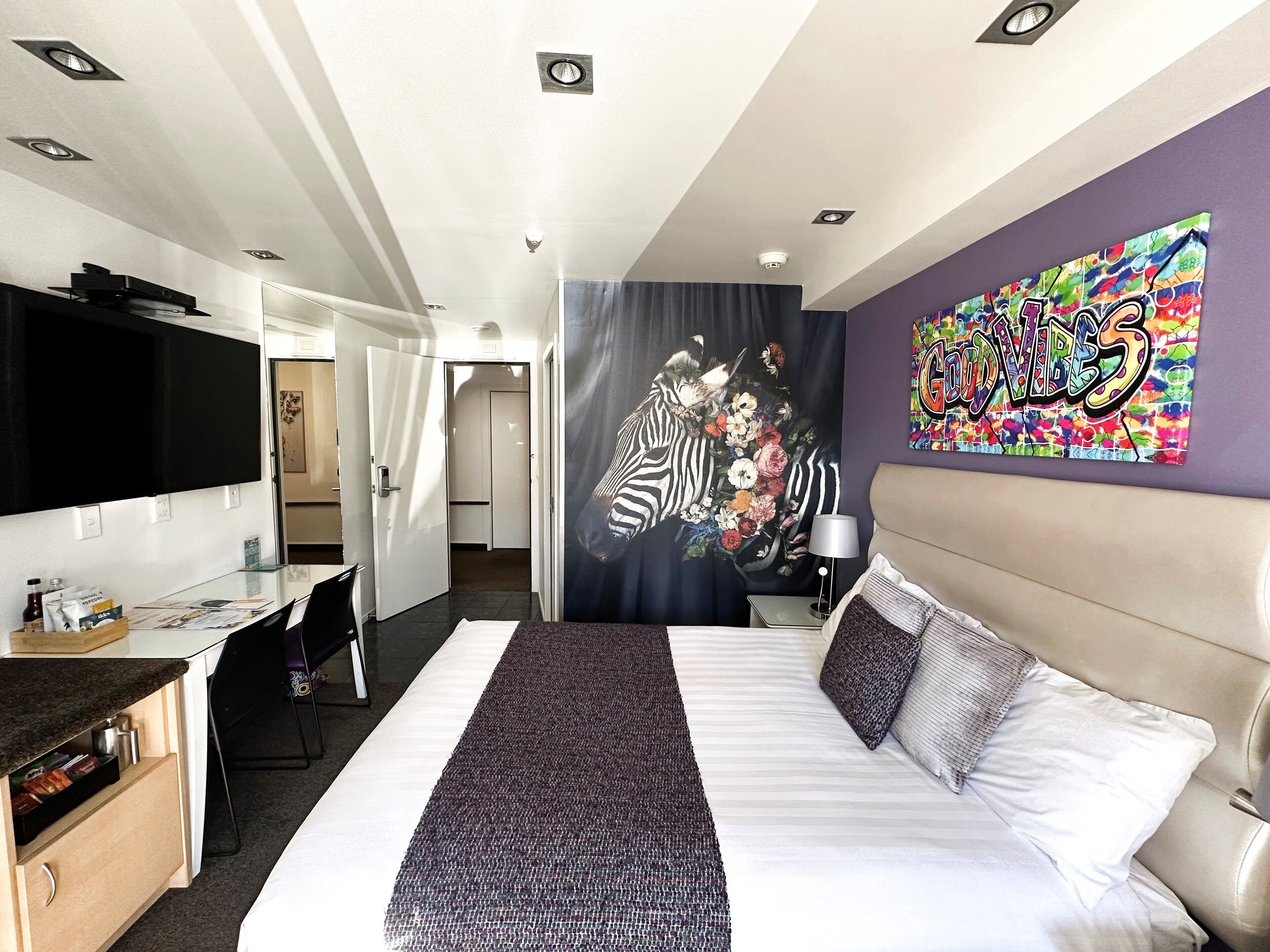 Quarto de hotel com cama, escrivaninha, televisão, quadro e porta do banheiro.