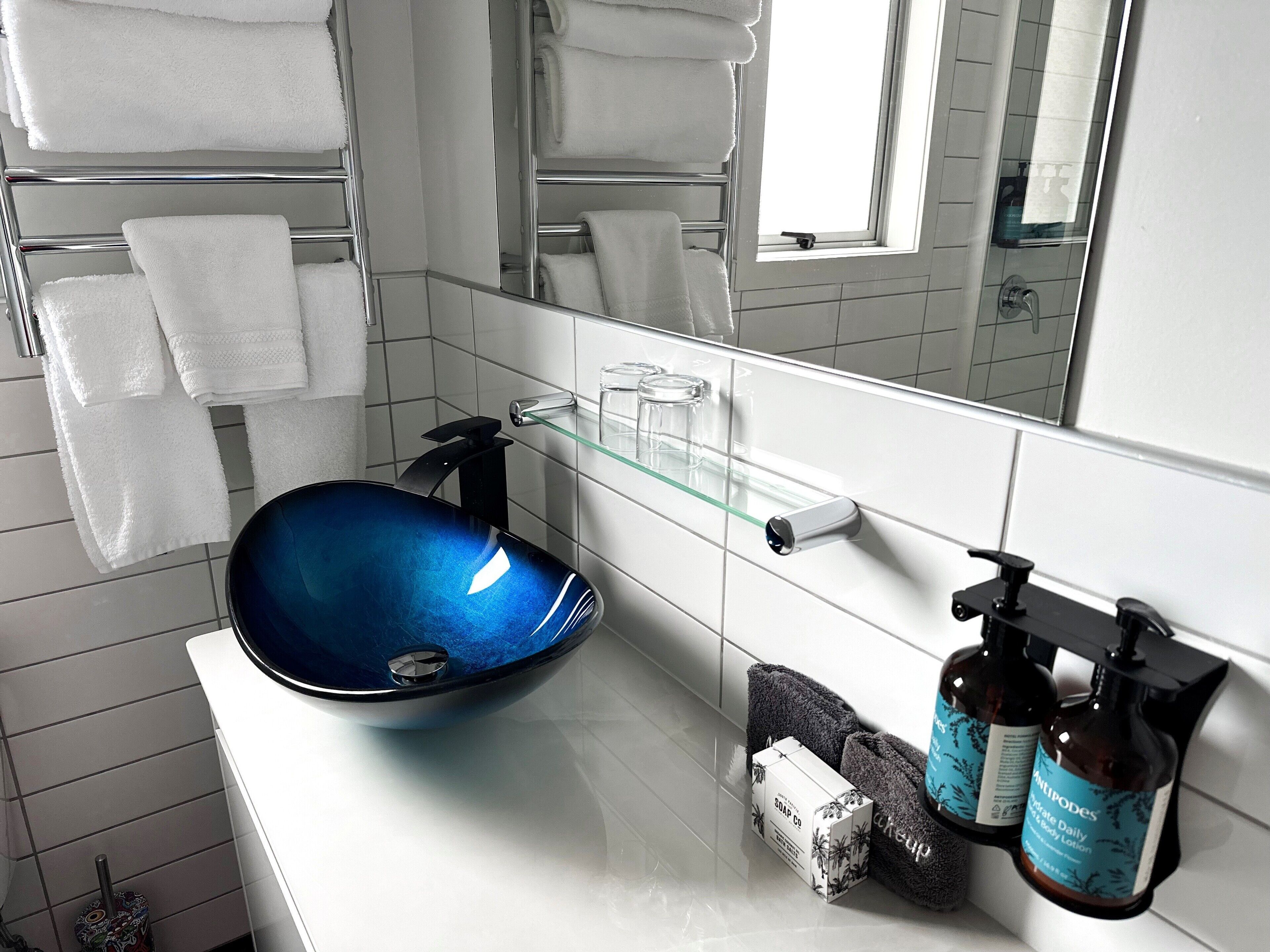 A modern bathroom with a blue glass sink, white tiled walls, a glass shelf, and neatly arranged towels.
