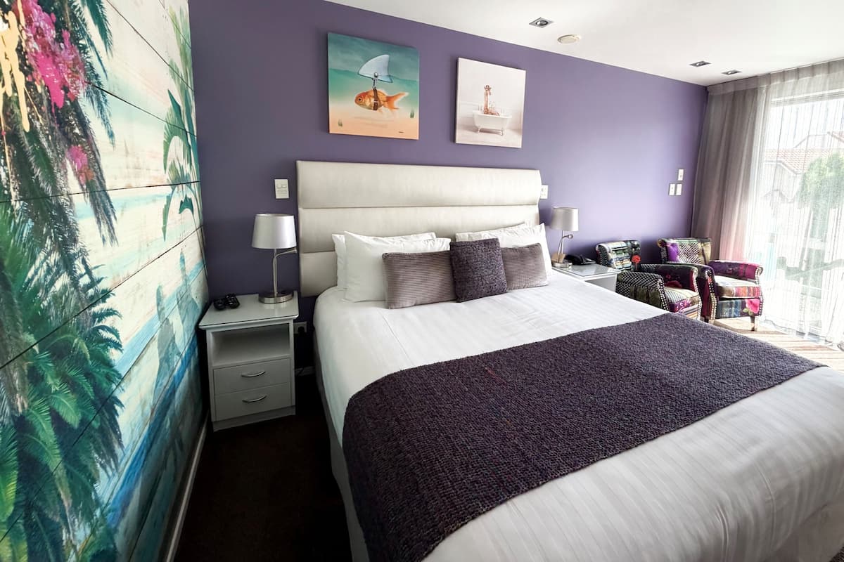 A hotel room with a large bed, a nightstand with a lamp, a painting on the wall, and a window with curtains.